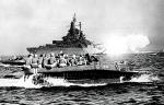 Amerykańska łódź desantowa płynie ku Okinawie, w tle pancernik USS „Tennessee” ostrzeliwuje wyspę, 1 kwietnia 1945 r. 