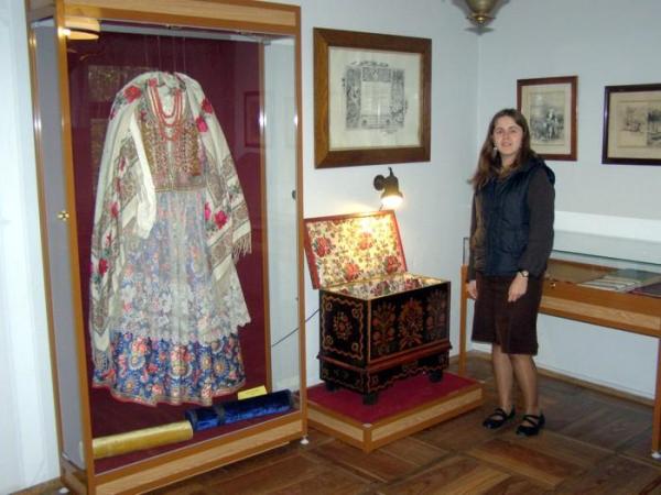 Pani Małgorzata Socha prezentuje strój krakowski uszyty przez żony Lucjana Rydal i Włodzimierza Tetmajera. Obok skrzynia w której strój dostarczono. Wykonaną jest z kolorowych kawałków drewna.